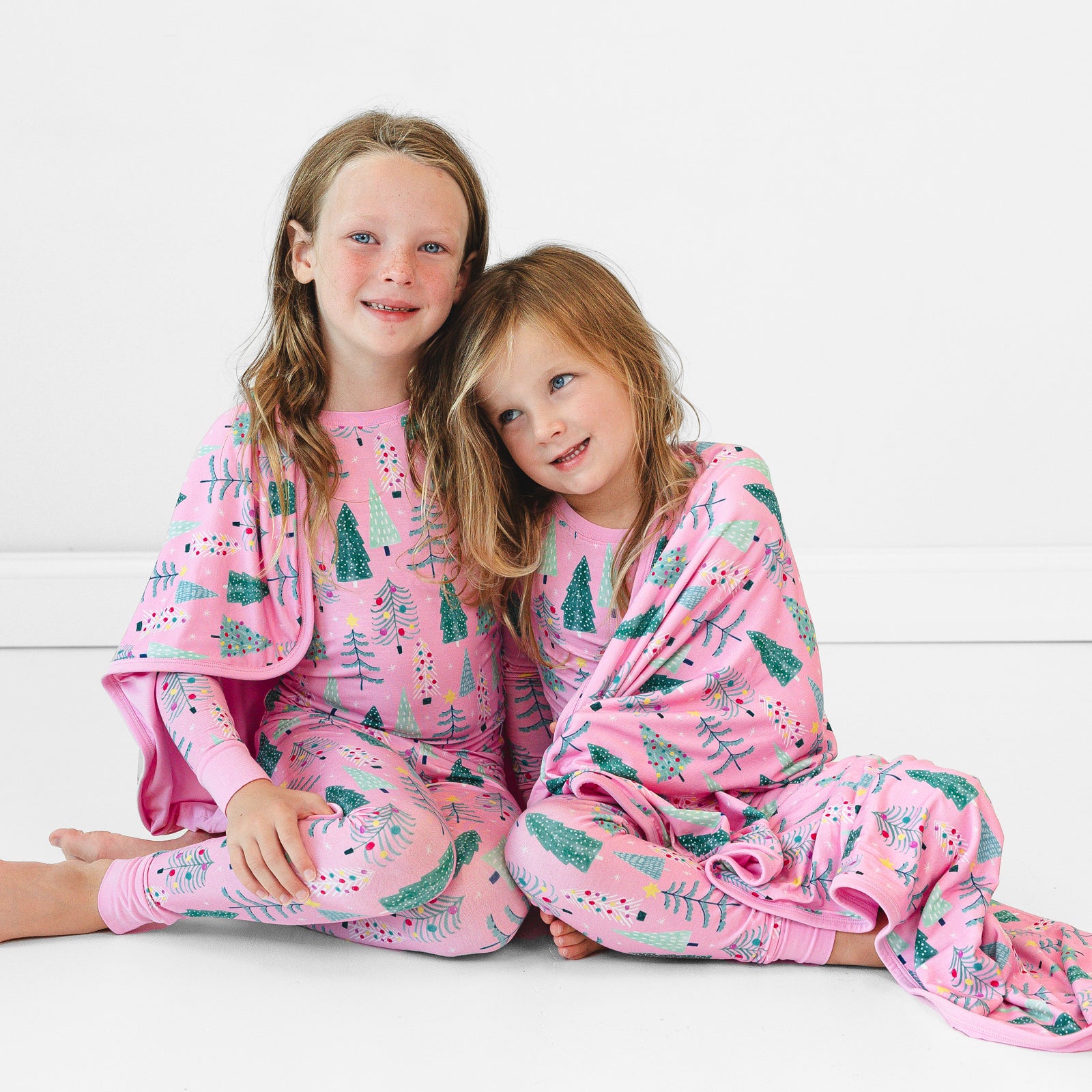 Two children sitting on the ground with a Pink Twinkling Trees Large Cloud Blanket draped over them and wearing matching pajamas