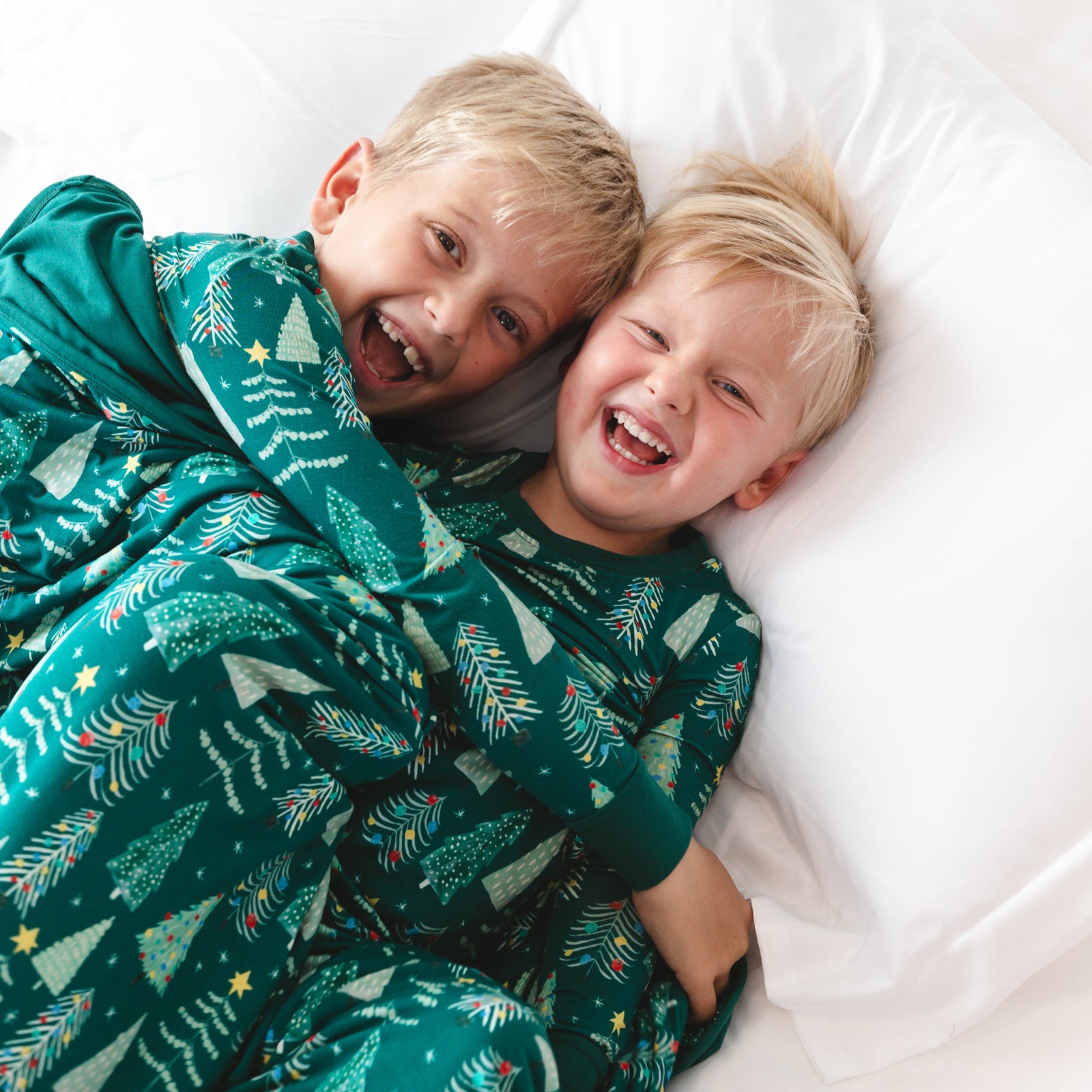 Top view image of two smiling children wrapped in the Green Twinkling Trees Large Cloud Blanket® while wearing the Green Twinkling Trees Two-piece Pajama Set