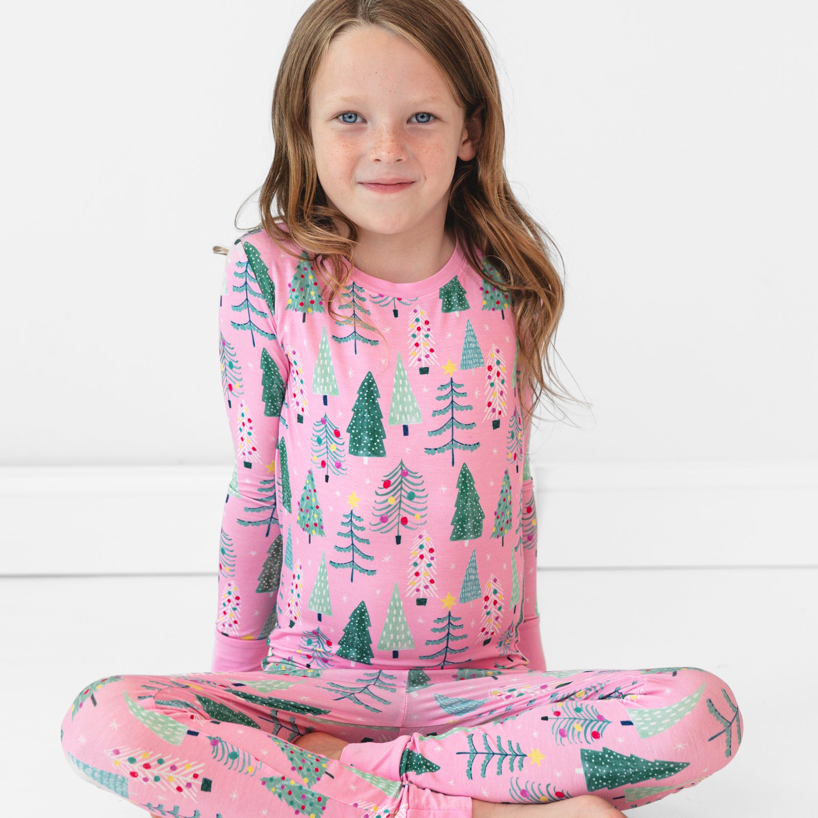 Image of a child sitting on the ground wearing a Pink Twinkling Trees Two-Piece Pajama Set