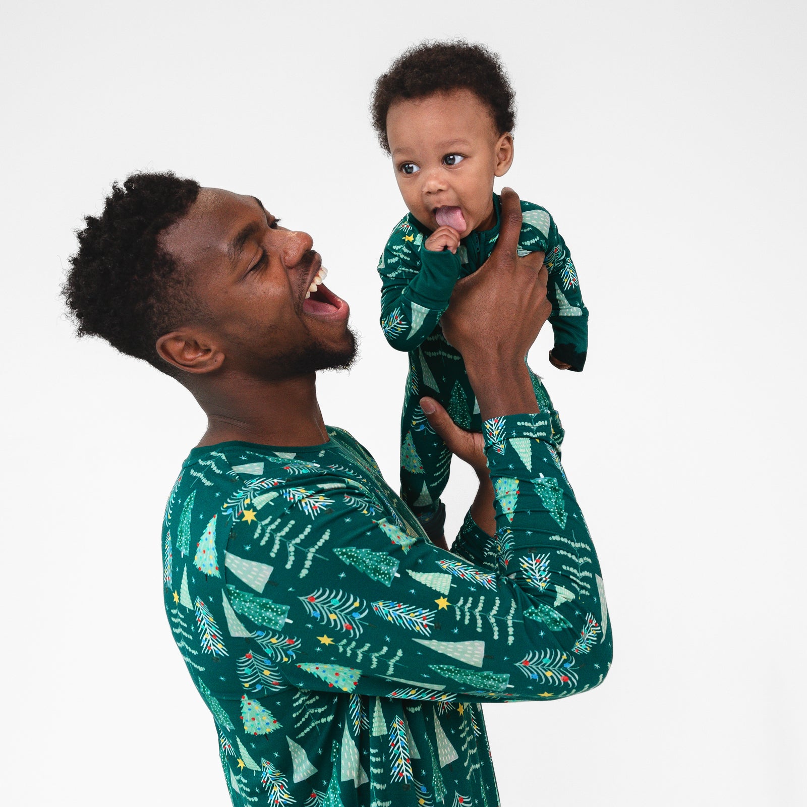 Male model is holding baby while wearing the Green Twinkling Trees Men's Pajama Top. Baby is in the Green Twinkling Trees Zippy