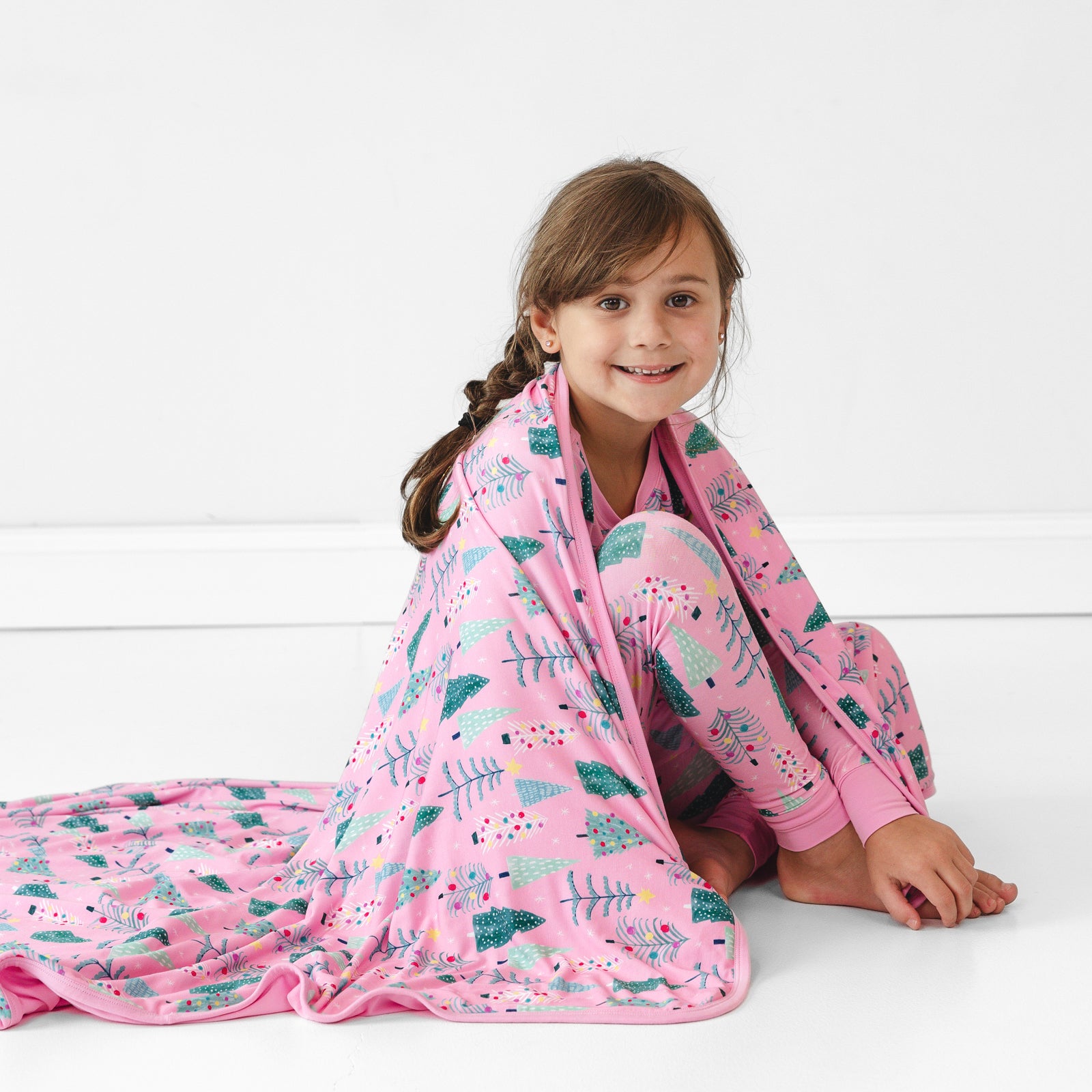 Child sitting on the ground with a Pink Twinkling Trees Large Cloud Blanket draped over them and wearing matching pajamas