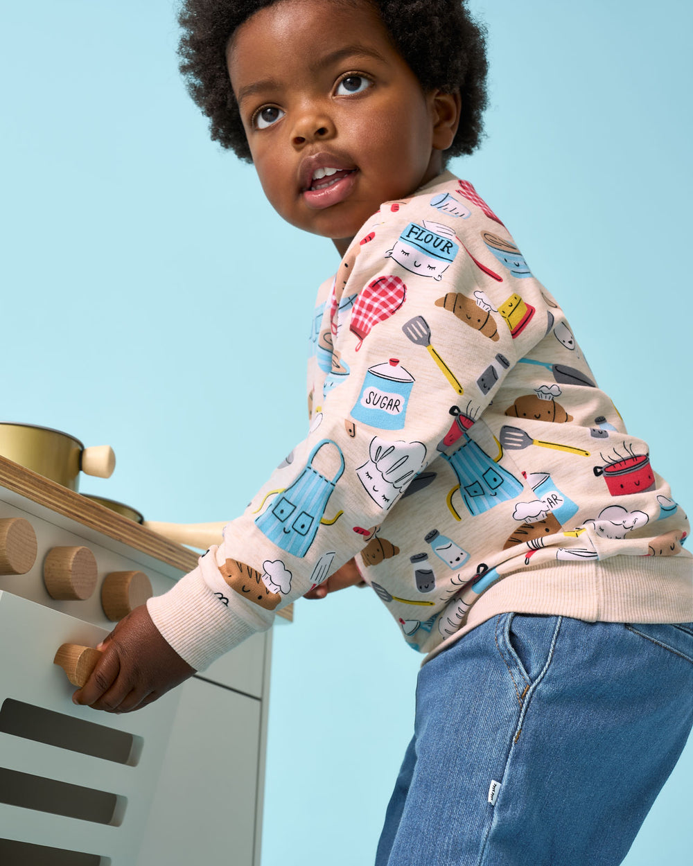 Child wearing the Little Chef Crewneck Sweatshirt playing with a wooden kitchen set