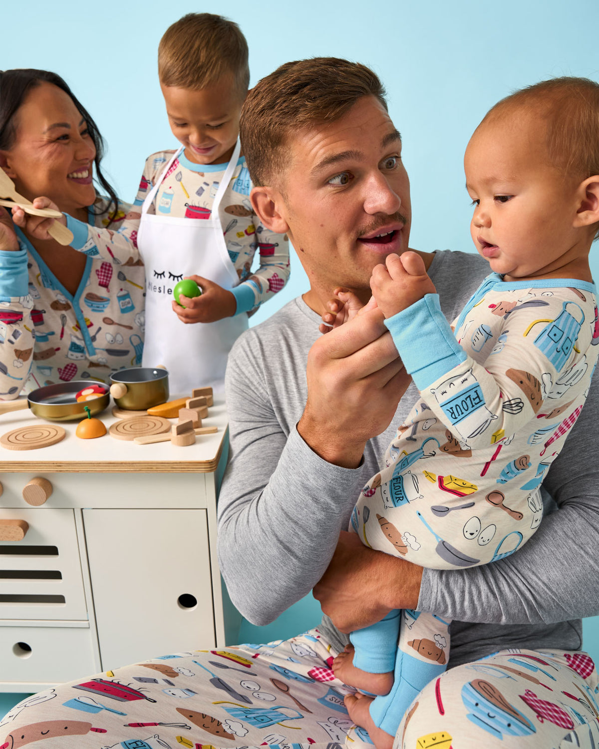Family of four wearing Little Chef pajamas playing with a wooden kitchen set
