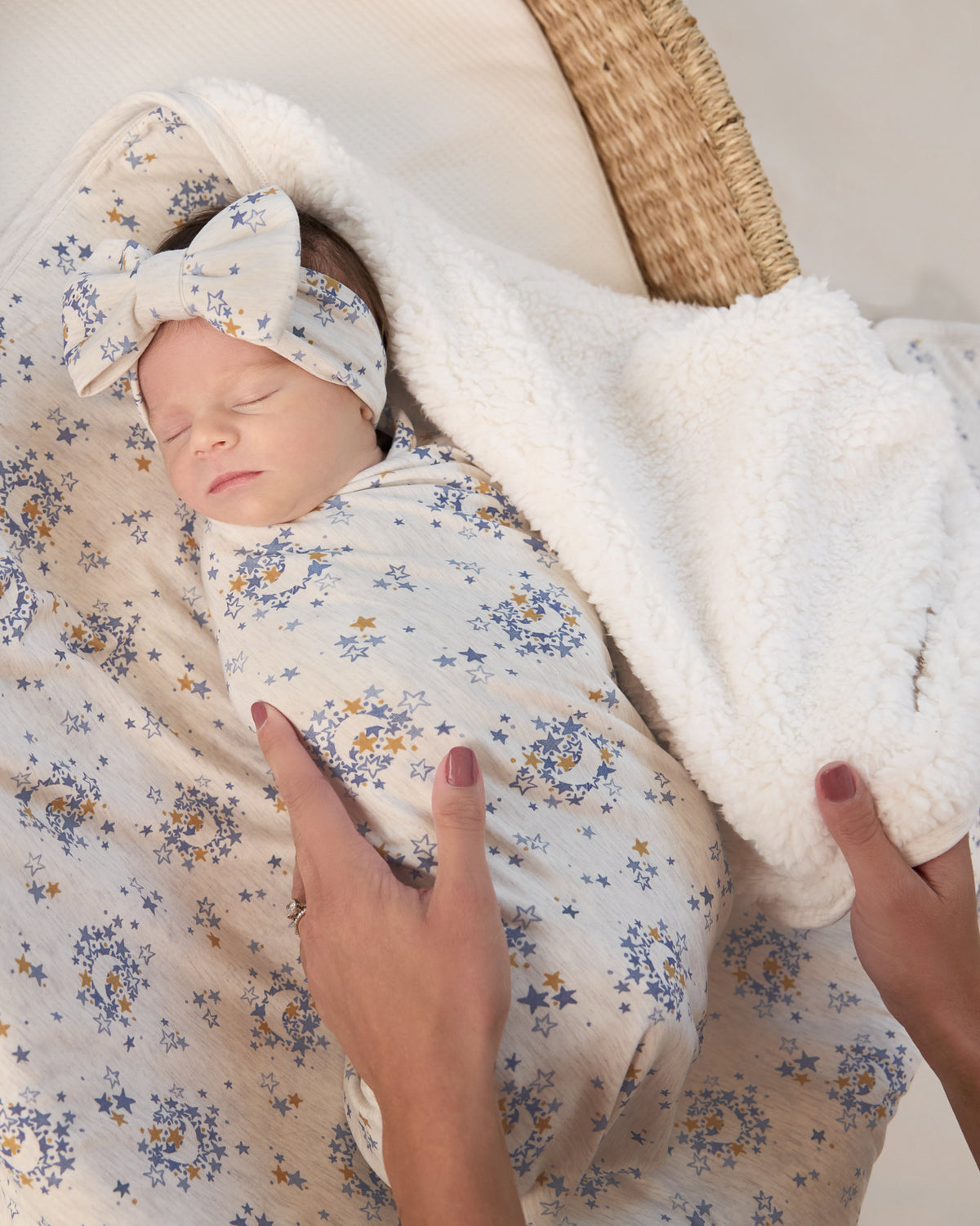 Newborn wearing the Lullaby Sky Swaddle & Luxe Bow Headband Set in a woven basket