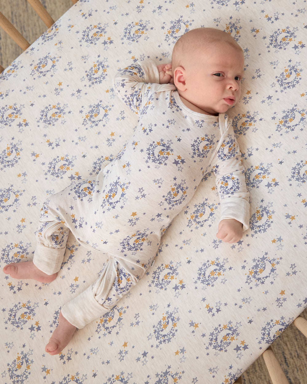 Aerial view of baby wearing the Lullaby Sky Crescent Zippy on a matching crib sheet