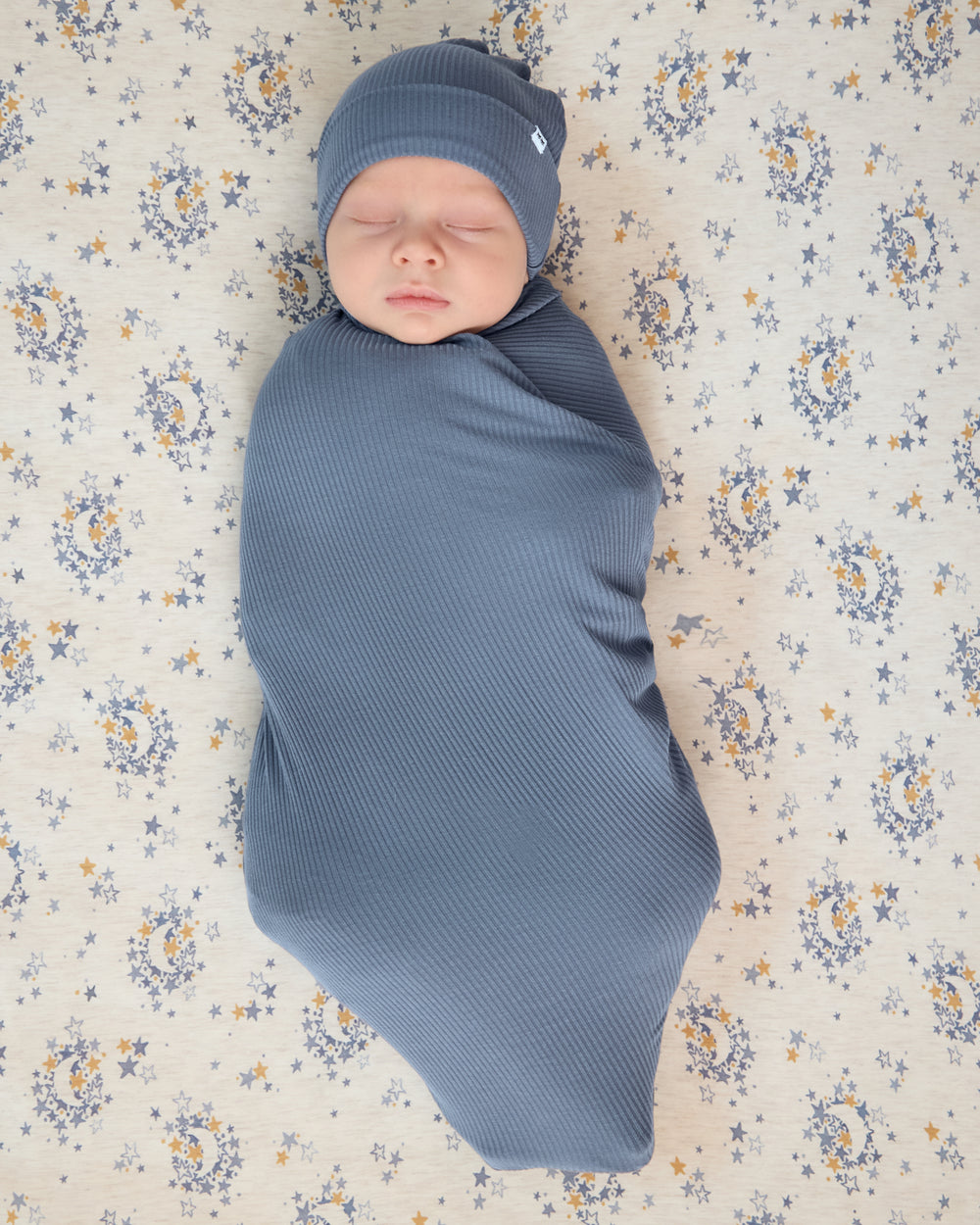 Newborn in a blue swaddle laying on the Lullaby Sky Fitted Crib Sheet