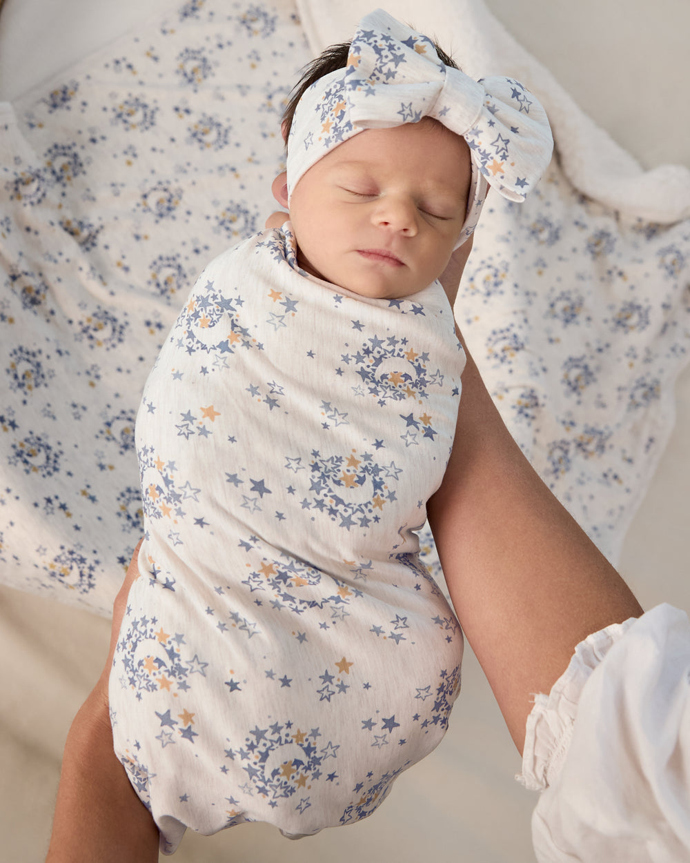 Adult holding newborn wrapped in the Lullaby Sky Swaddle & Luxe Bow Headband Set