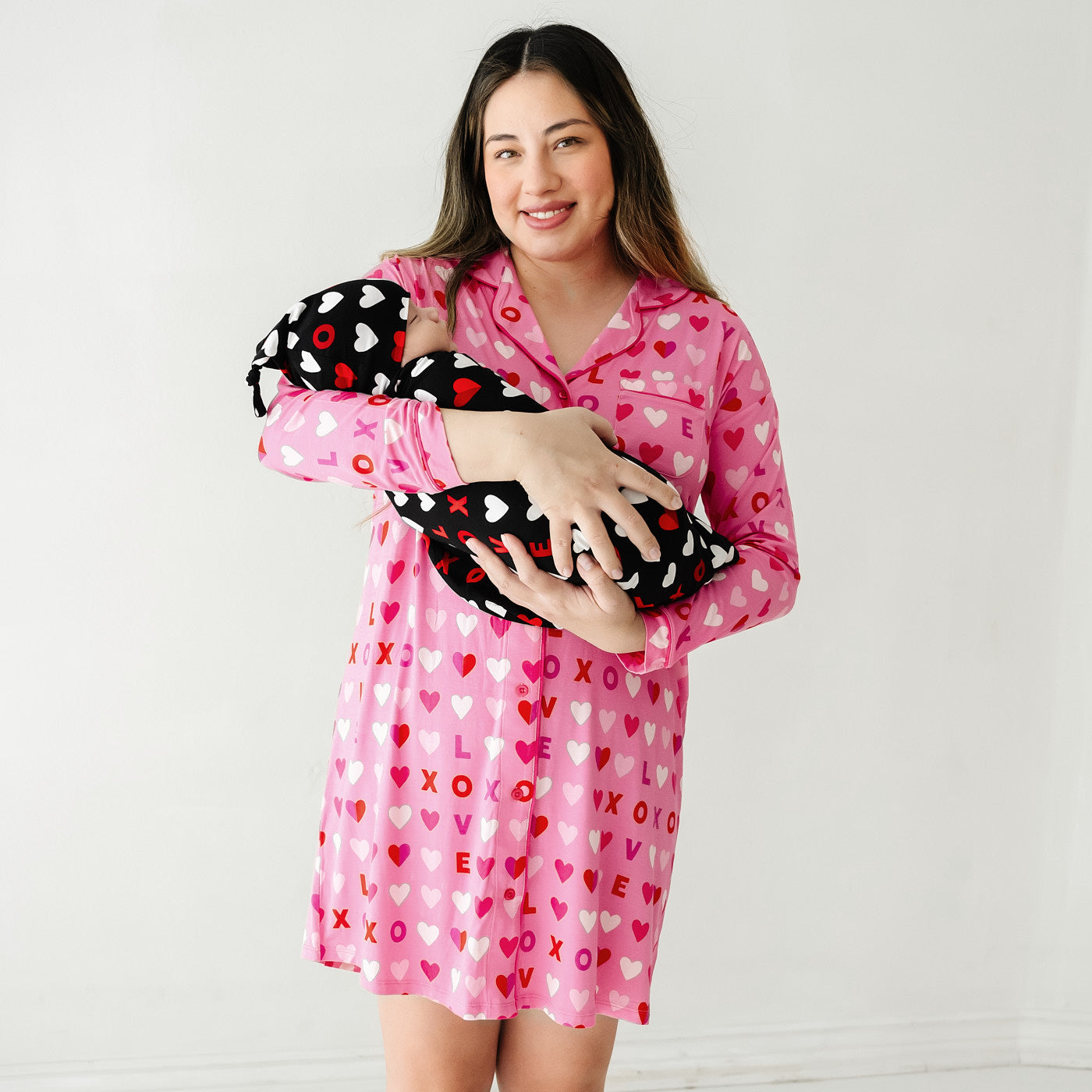 Mom holding her child swaddled in a Black XOXO swaddle and hat set. Mom is wearing a women's Pink XOXO long sleeve sleep shirt