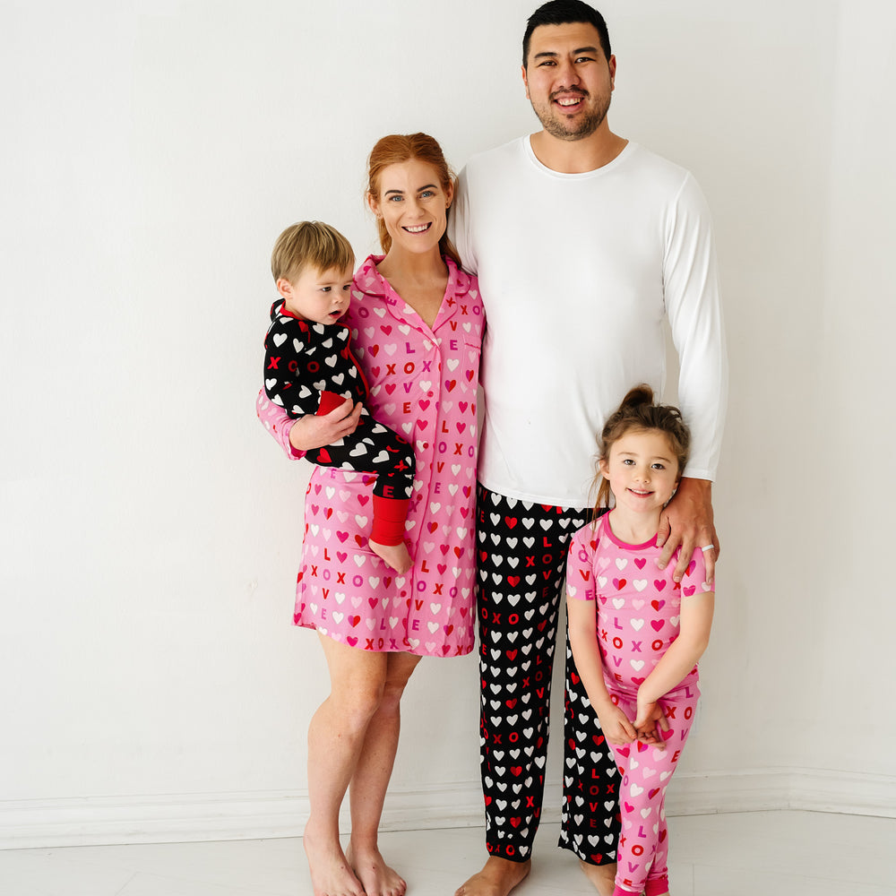 Click to see full screen - Family of four wearing coordinating XOXO pajama sets. Dad is wearing men's Black XOXO pajama pants paired with a men's solid white pajama top. Mom is wearing Pink XOXO women's long sleeve sleep shirt. Kids are wearing Black and Pink XOXO pajamas in two piece and zippy styles.