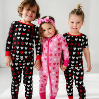 Three children holding hands wearing coordinating XOXO pajama sets. One is wearing a Pink XOXO zippy and the other two are wearing Black XOXO pjs in long sleeve and short sleeve two piece styles
