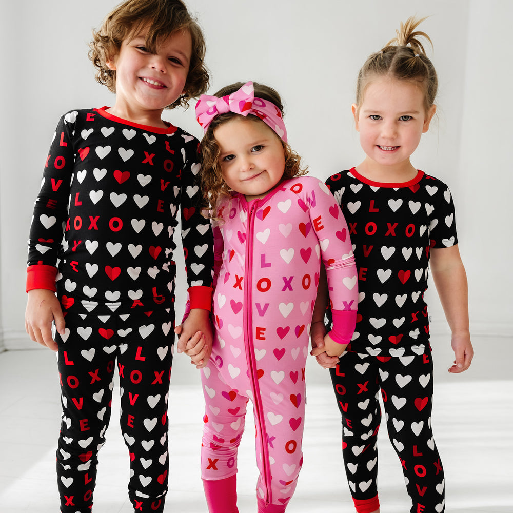 Click to see full screen - Three children holding hands wearing coordinating XOXO pajama sets. One is wearing a Pink XOXO zippy and the other two are wearing Black XOXO pjs in long sleeve and short sleeve two piece styles
