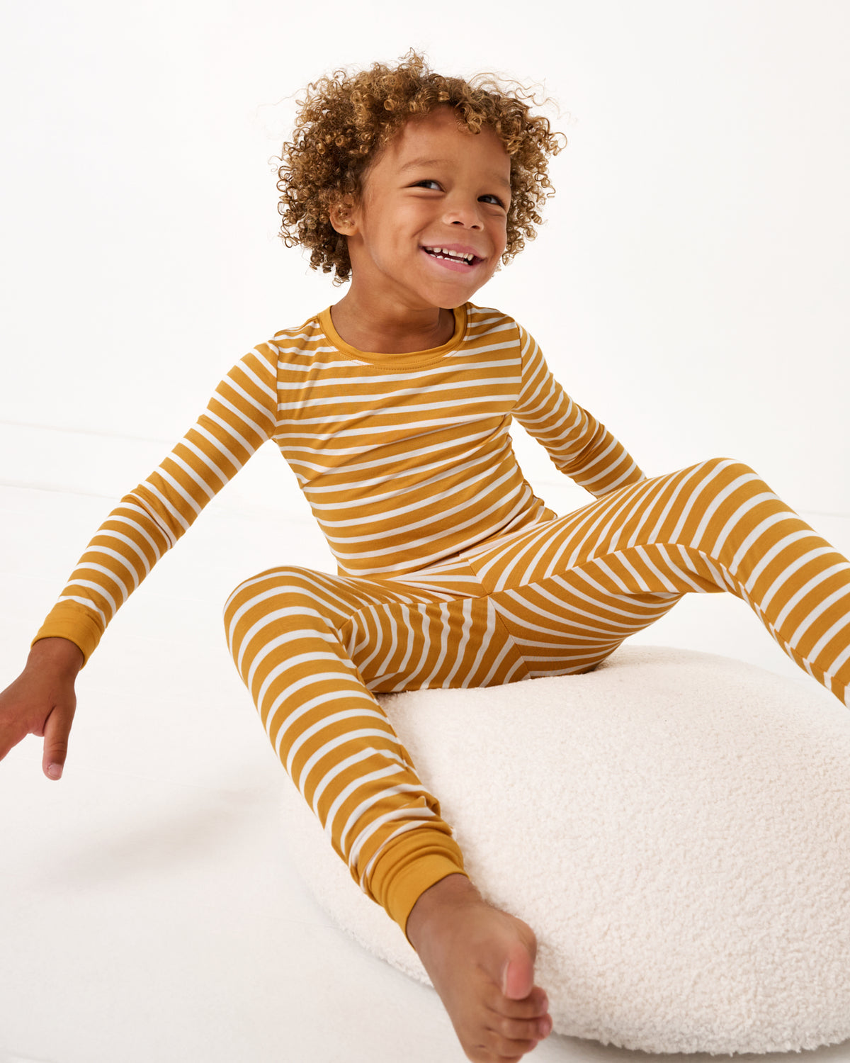 Boy sitting on a boucle ottoman wearing the Marigold Stripe Two-Piece Pajama Set