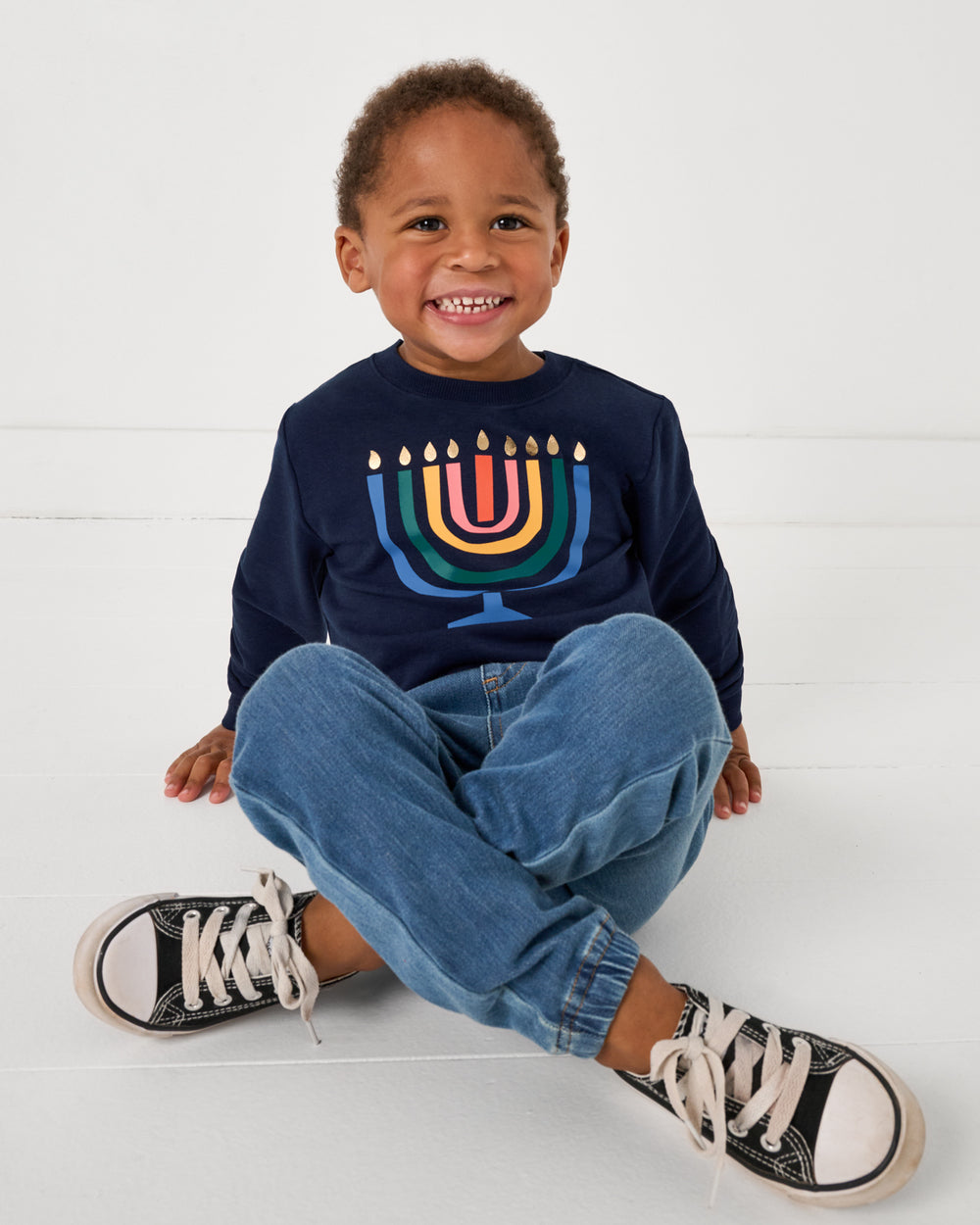 Sitting boy wearing the Menorah Crewneck Sweatshirt with denim joggers