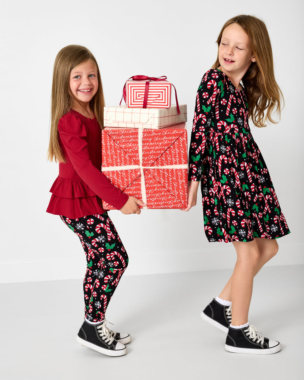 Two girls in festive outfits with candy cane patterns holding gift boxes.