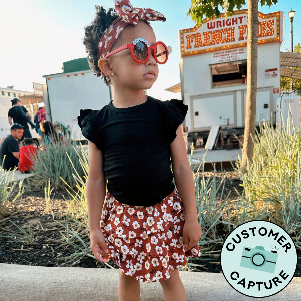 Customer Capture image of a child wearing a Mocha Blossom printed ruffle skort paired with a Black flutter sleeve tee and Mocha Blossom luxe bow headband