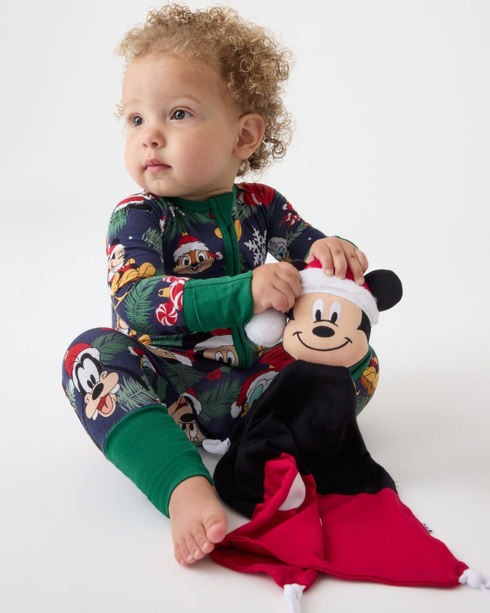 A toddler sits on a white studio floor wearing a festive Christmas-themed Mickey Mouse pajama set. The one-piece PJ features a dark navy blue background with green cuffs, a green zipper, and a holiday print of Mickey, Minnie, and other Disney characters.