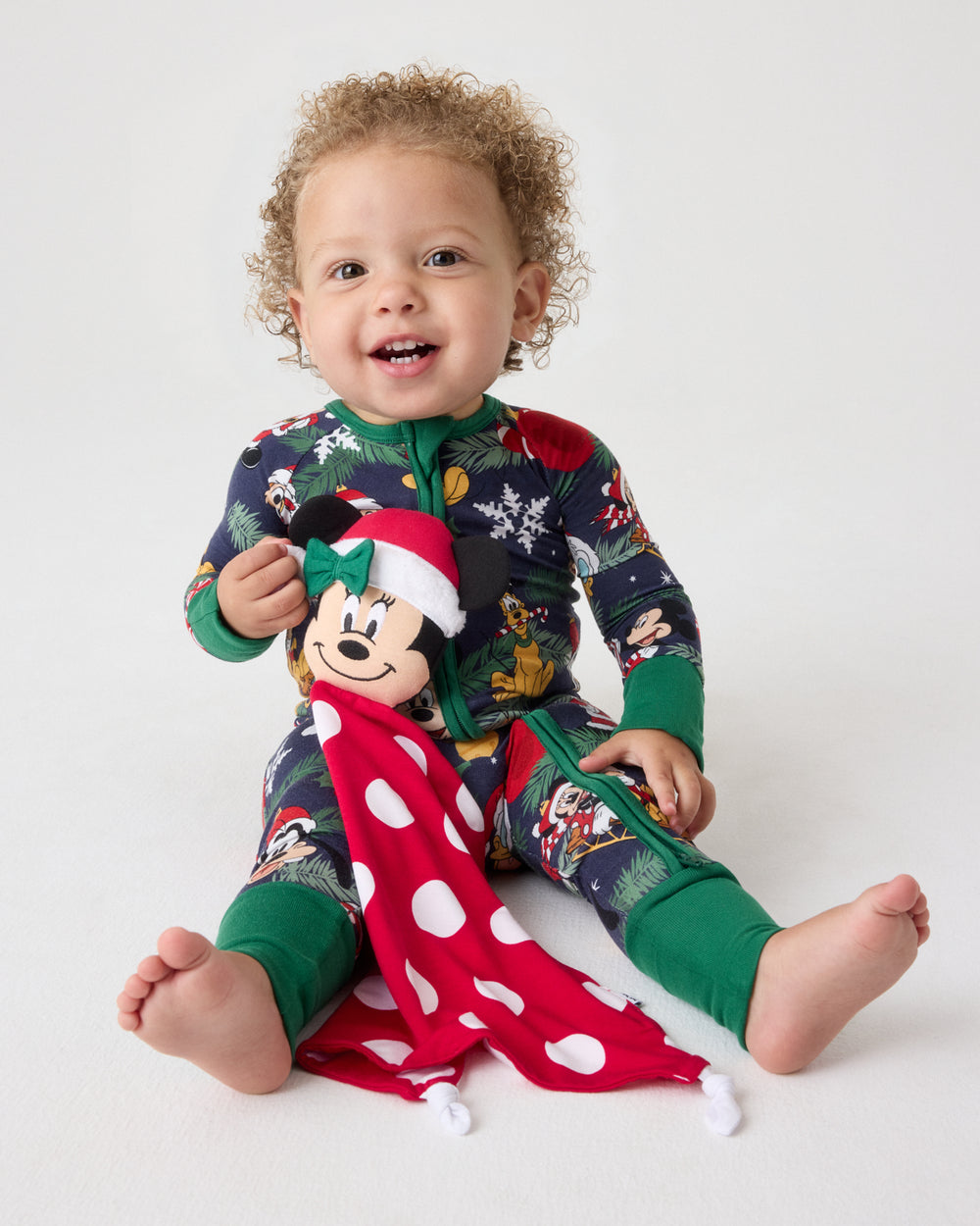 A smiling toddler sits on a light gray studio background wearing a festive Christmas-themed long-sleeve pajama set with a colorful print and green cuffs. The child holds a plush toy with a Santa hat and a red-and-white polka dot scarf.
