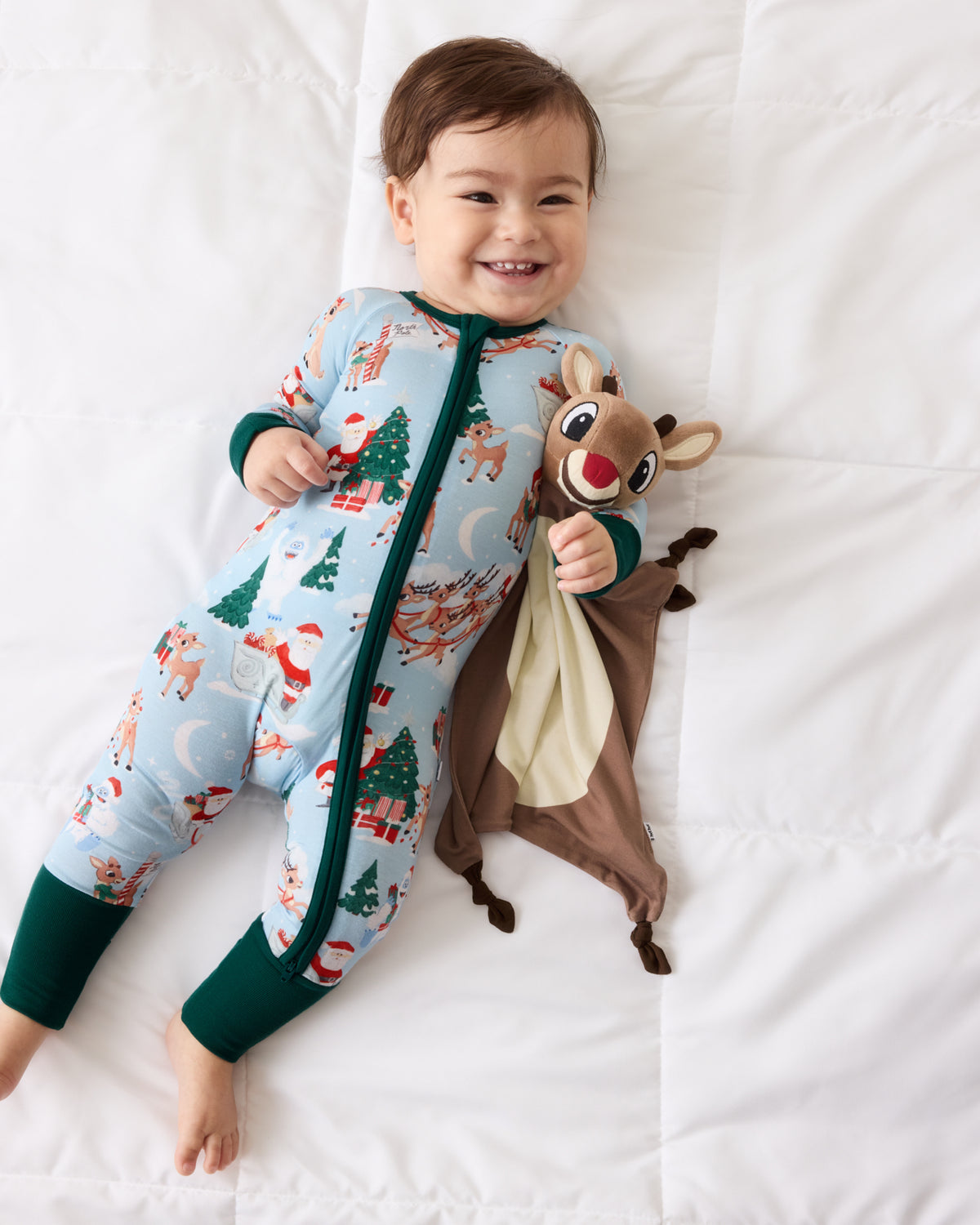 A baby lying on a white quilted blanket wearing a light blue Christmas-themed footed pajama with green trim and holding a brown reindeer plush toy.