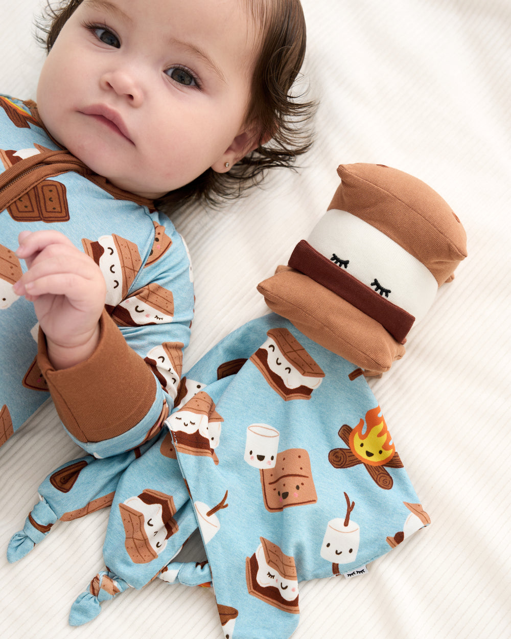 A baby lying on a white blanket wearing a light blue footed sleeper with a brown and white s'mores print, next to a matching plush s'mores comforter.
