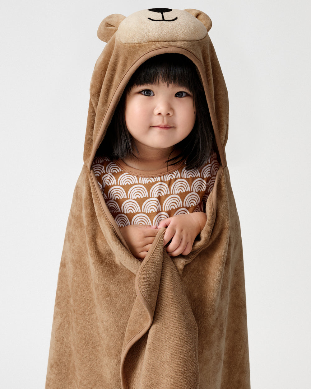 A young child wearing a brown plush hooded towel wrap with bear ears and a stitched bear face, over a patterned brown and white top, photographed against a plain light gray background.