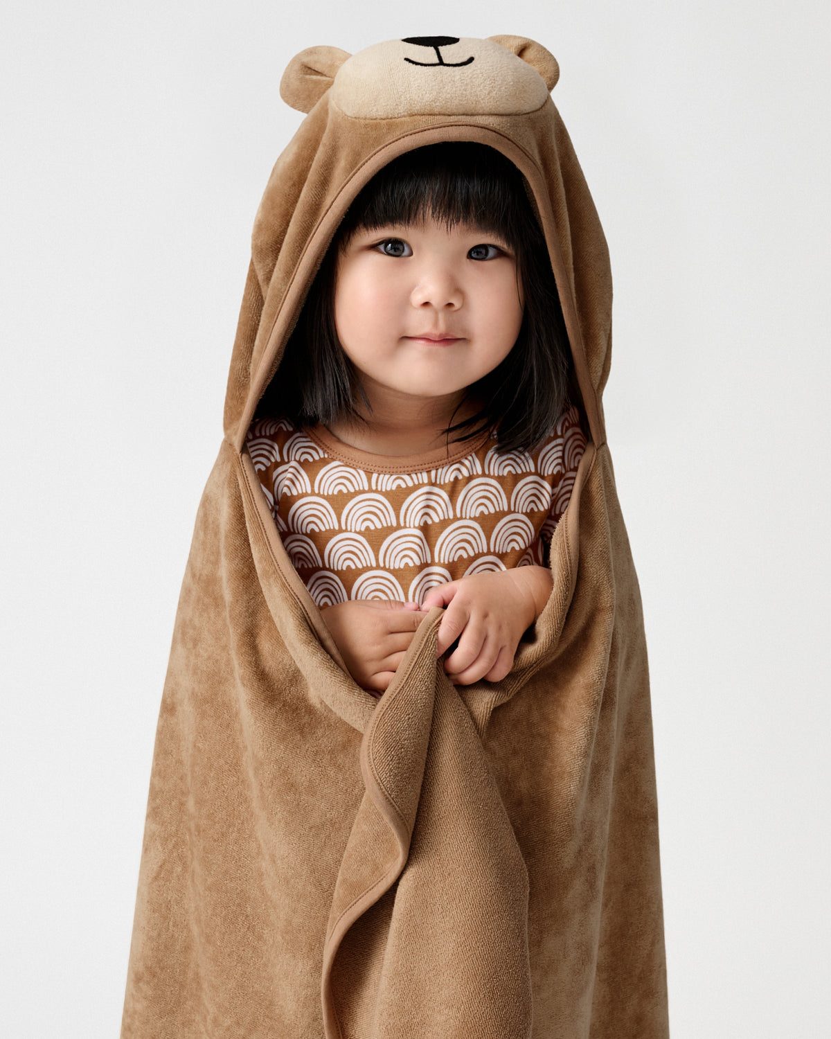A young child wrapped in a brown plush hooded towel with bear ears and a bear face on the hood, standing against a light gray studio background.