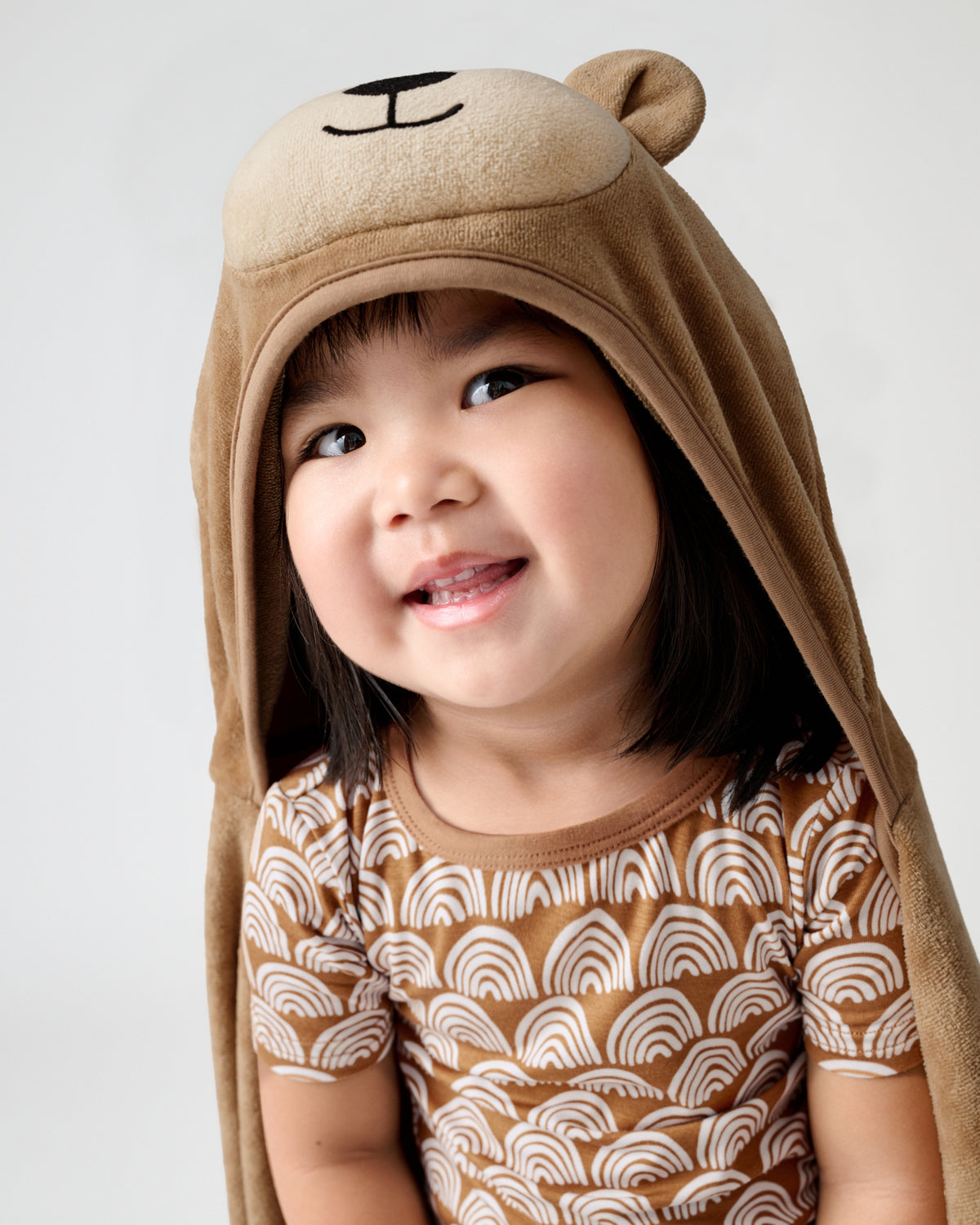 A smiling young child wearing a brown plush hooded towel or cape with bear ears and a bear face, paired with a brown and white patterned short-sleeve top, against a light gray studio background.