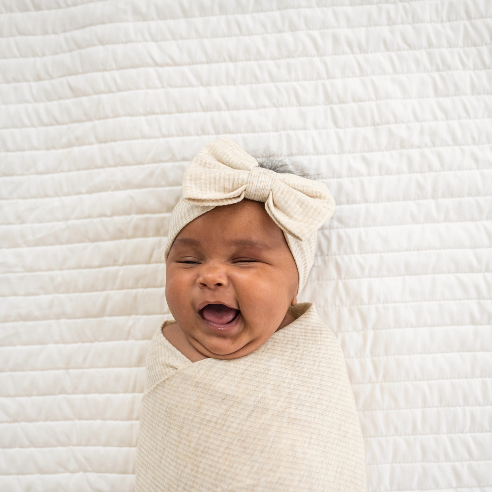 Close up immage of a child swaddled in a Heather Oatmeal Ribbed Swaddle and Luxe Bow Headband Set