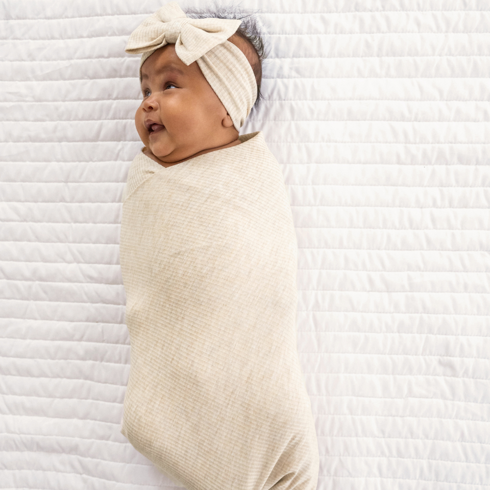 Image of a child swaddled in a Heather Oatmeal Ribbed Swaddle and Luxe Bow Headband Set