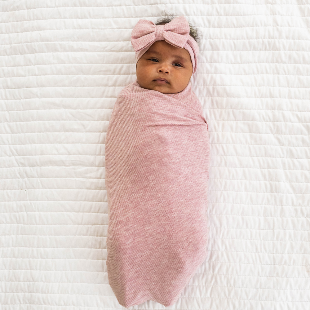 Child on a bed swaddled in a Heather Mauve Ribbed Swaddle and luxe bow Headband set