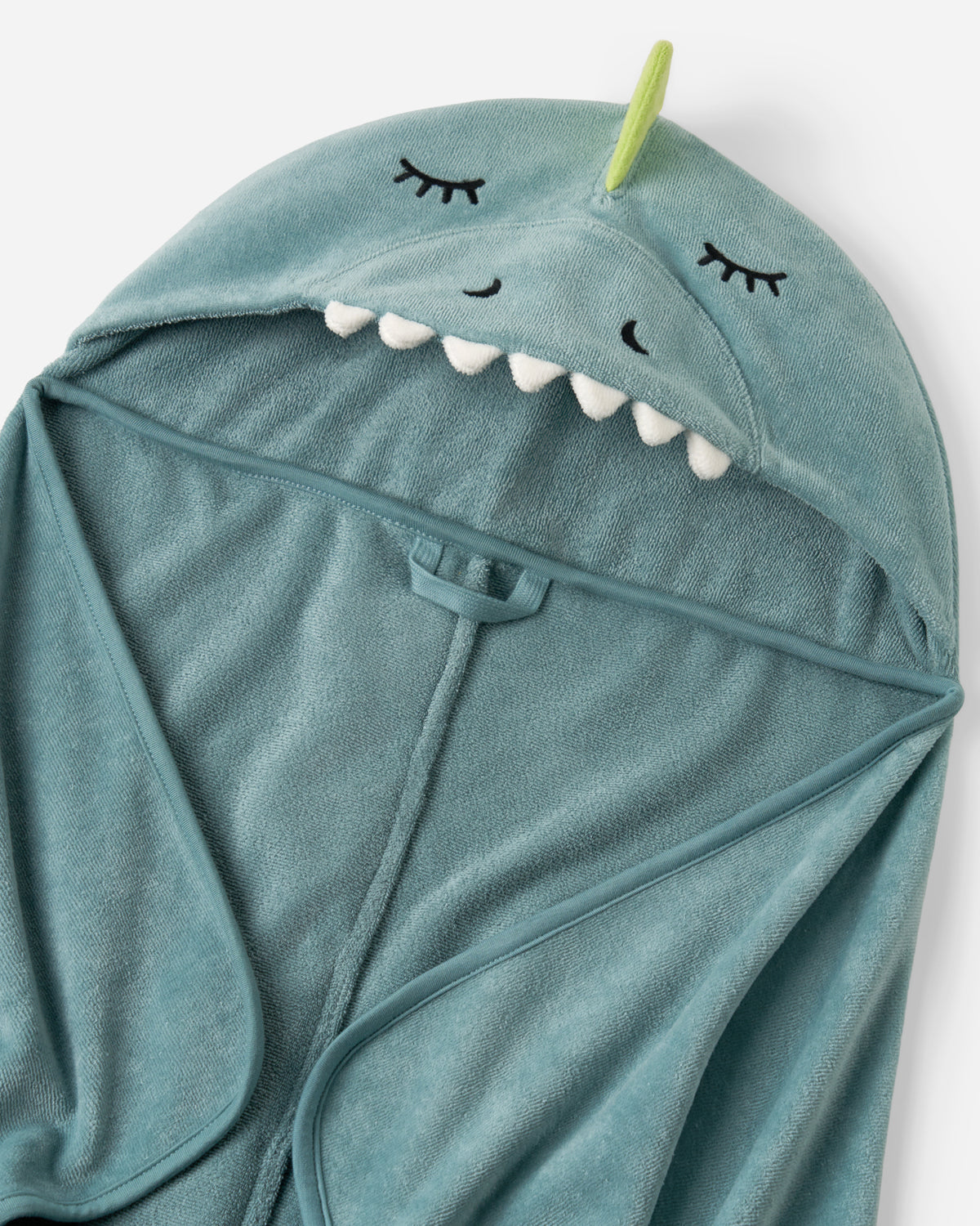 A close-up of a teal blue dinosaur-themed hooded towel or robe, featuring embroidered closed eyes, white teeth, and a small green horn on a light gray background.