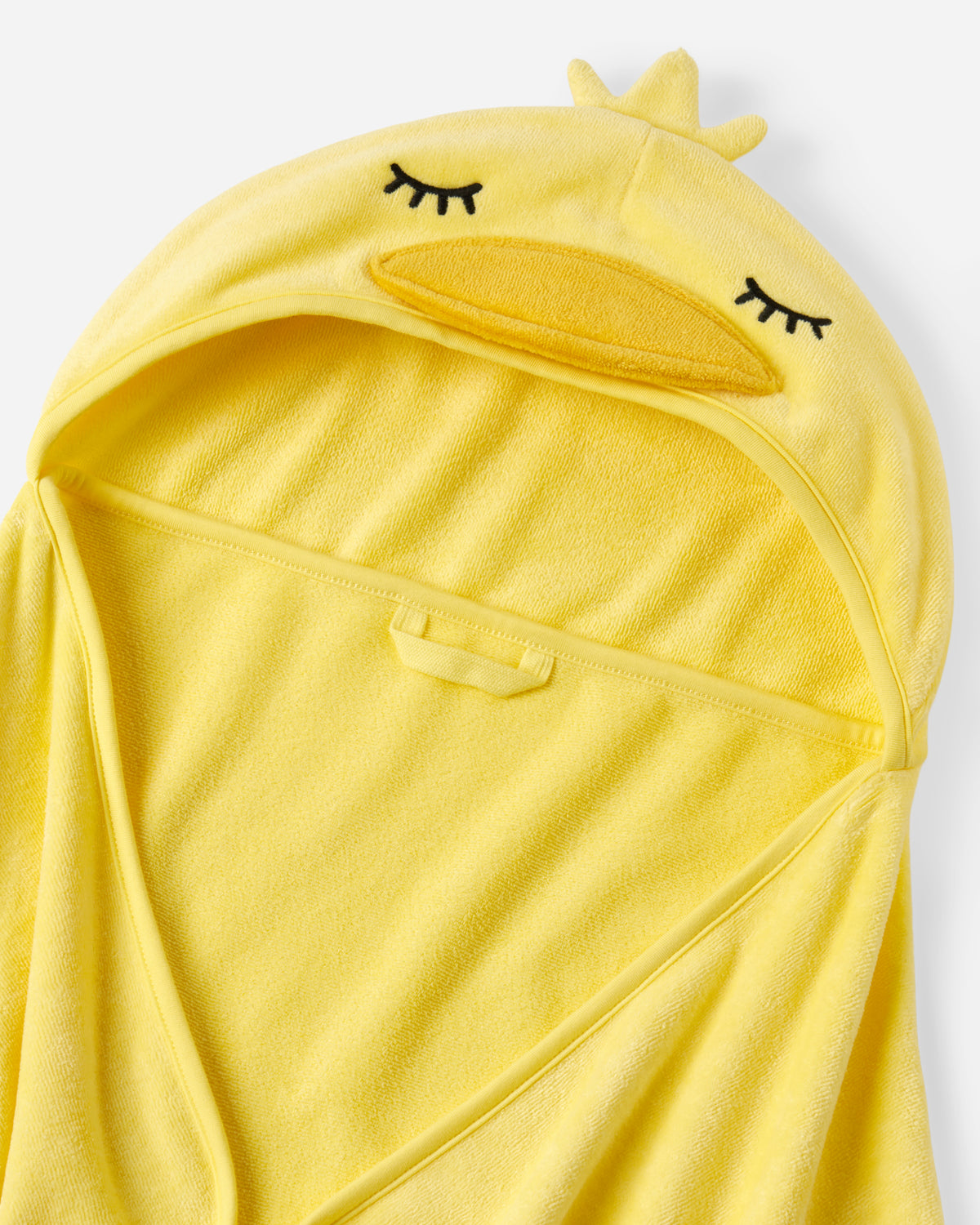 A close-up of a yellow hooded towel or wrap with a duck-face design, including embroidered closed eyes, a beak, and a small crown on the hood, shown against a plain light background.