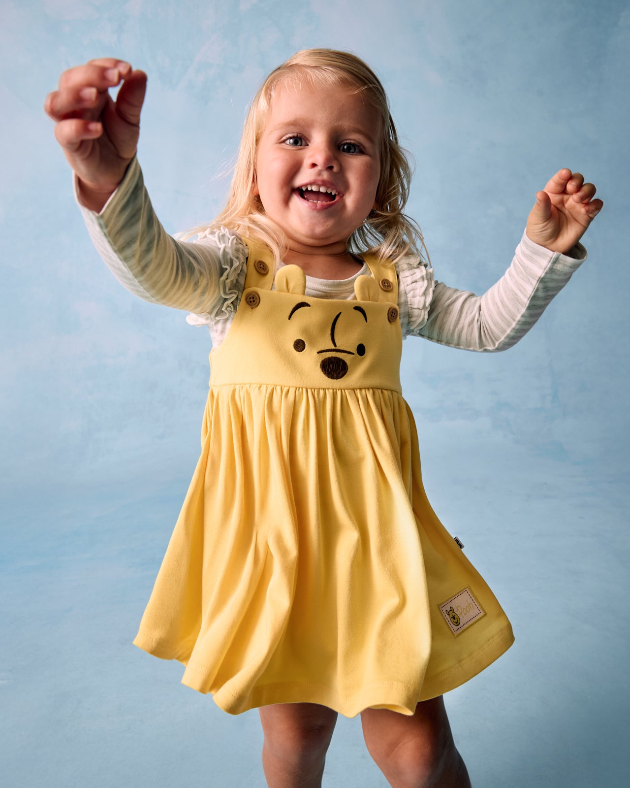 Girl wearing the Disney Winnie The Pooh Overall Dress layered over a flutter bodysuit