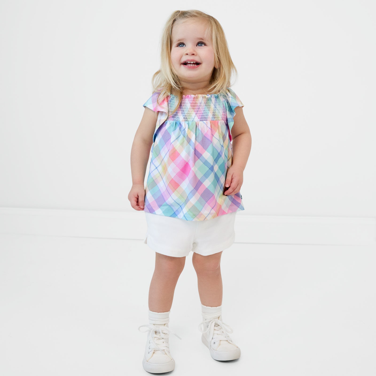 Standing child wearing the Pastel Plaid Smocked Flutter Top with bright white denim shorts