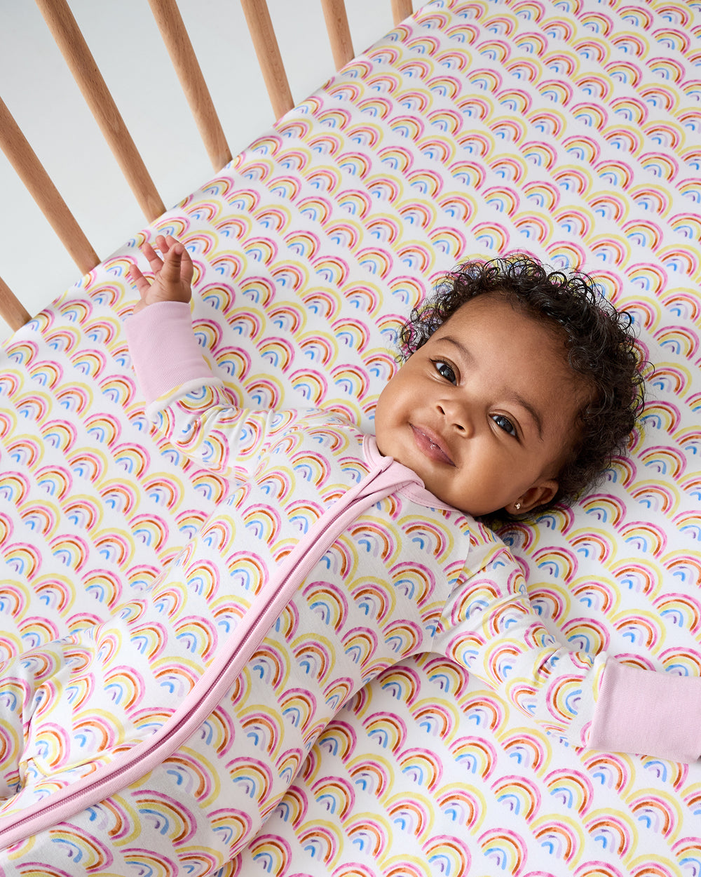 Baby laying on the Pastel Rainbows Fitted Crib Sheet wearing the matching zippy