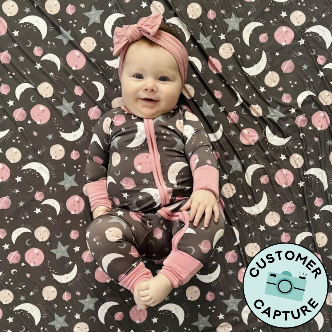 Customer Capture image of a child wearing a Pink to the moon and back zippy laying on a matching cloud blanket