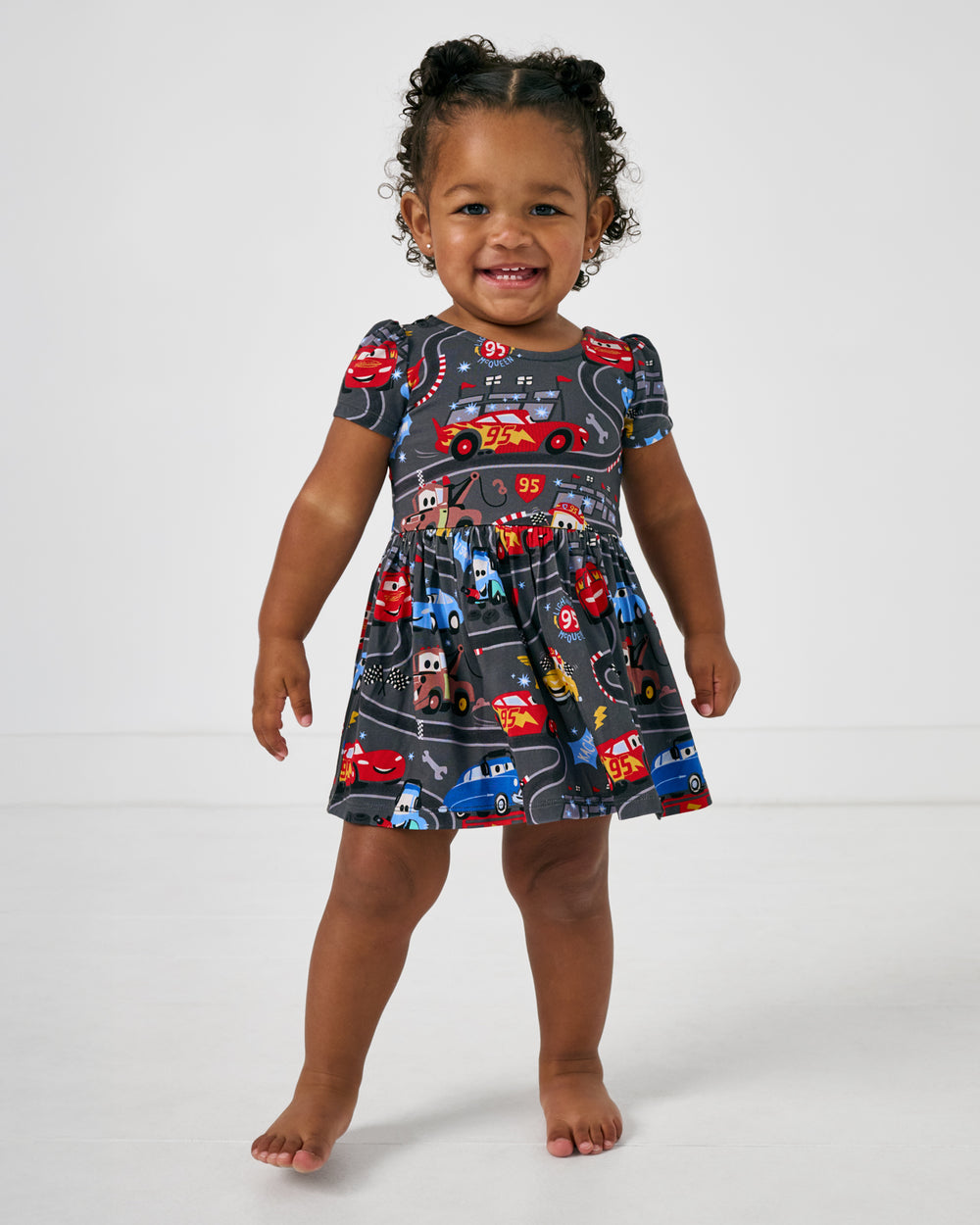 Standing toddler wearing the Disney & Pixar Piston Cup Bow Back Skater Dress with Bodysuit