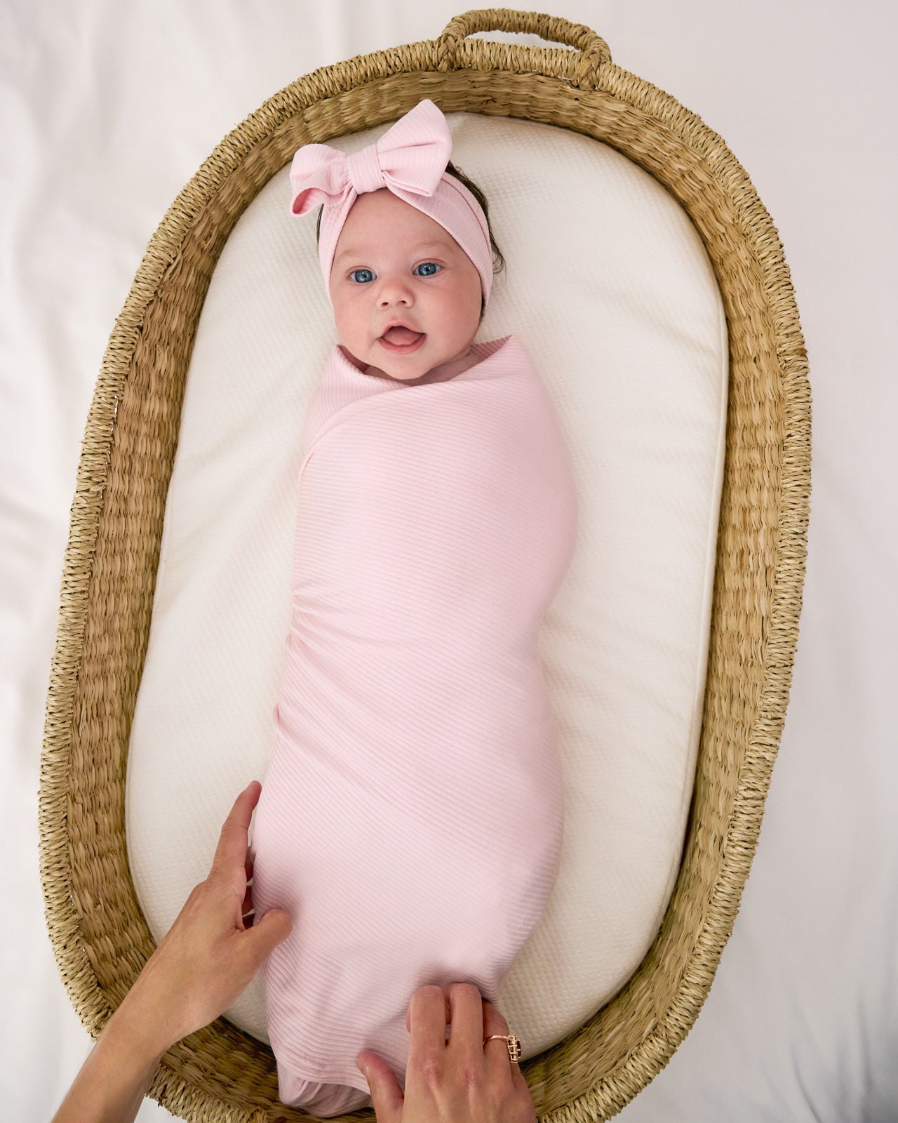 Baby in a basket crib wearing the Prima Pink Ribbed Swaddle & Luxe Bow Headband Set