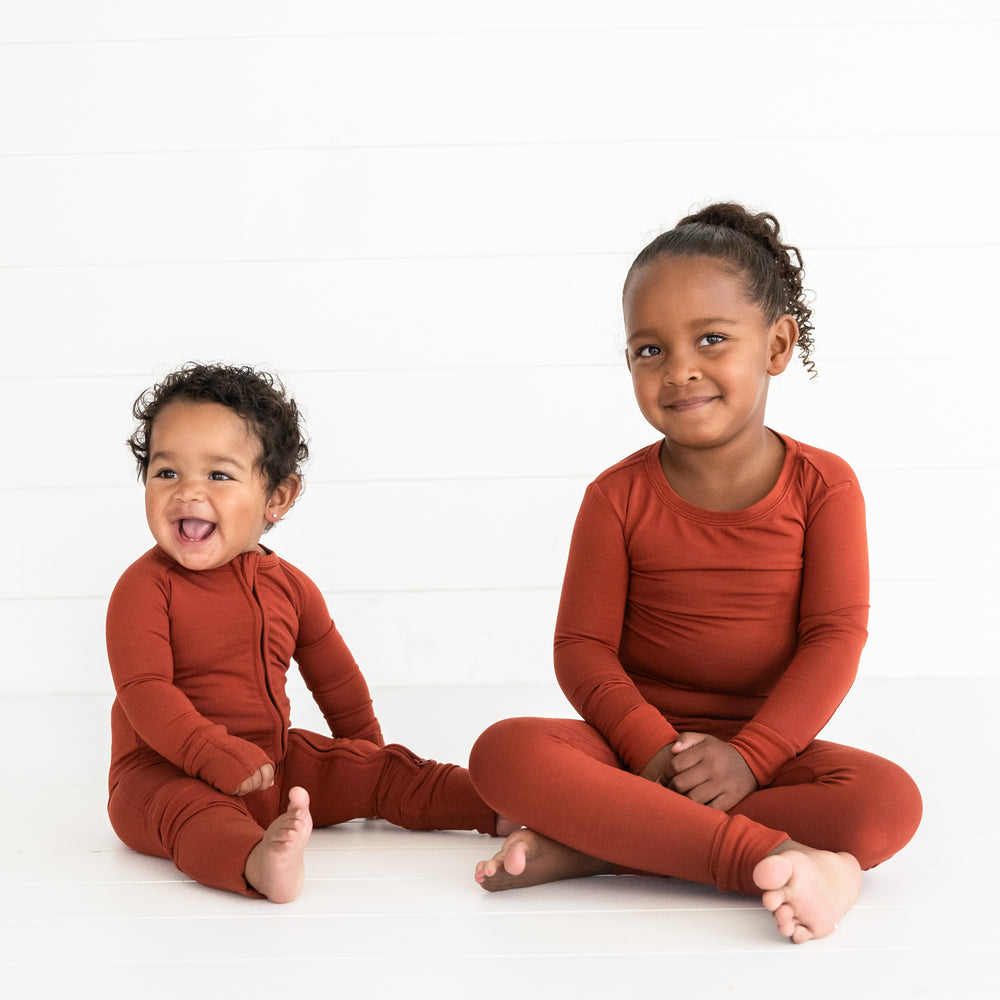 Two children sitting together wearing a Cozy Rust two piece pj set and a matching zippy