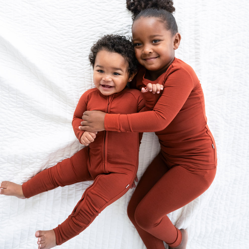 Two children cuddling on a bed wearing a Cozy Rust two piece pj set and a matching zippy