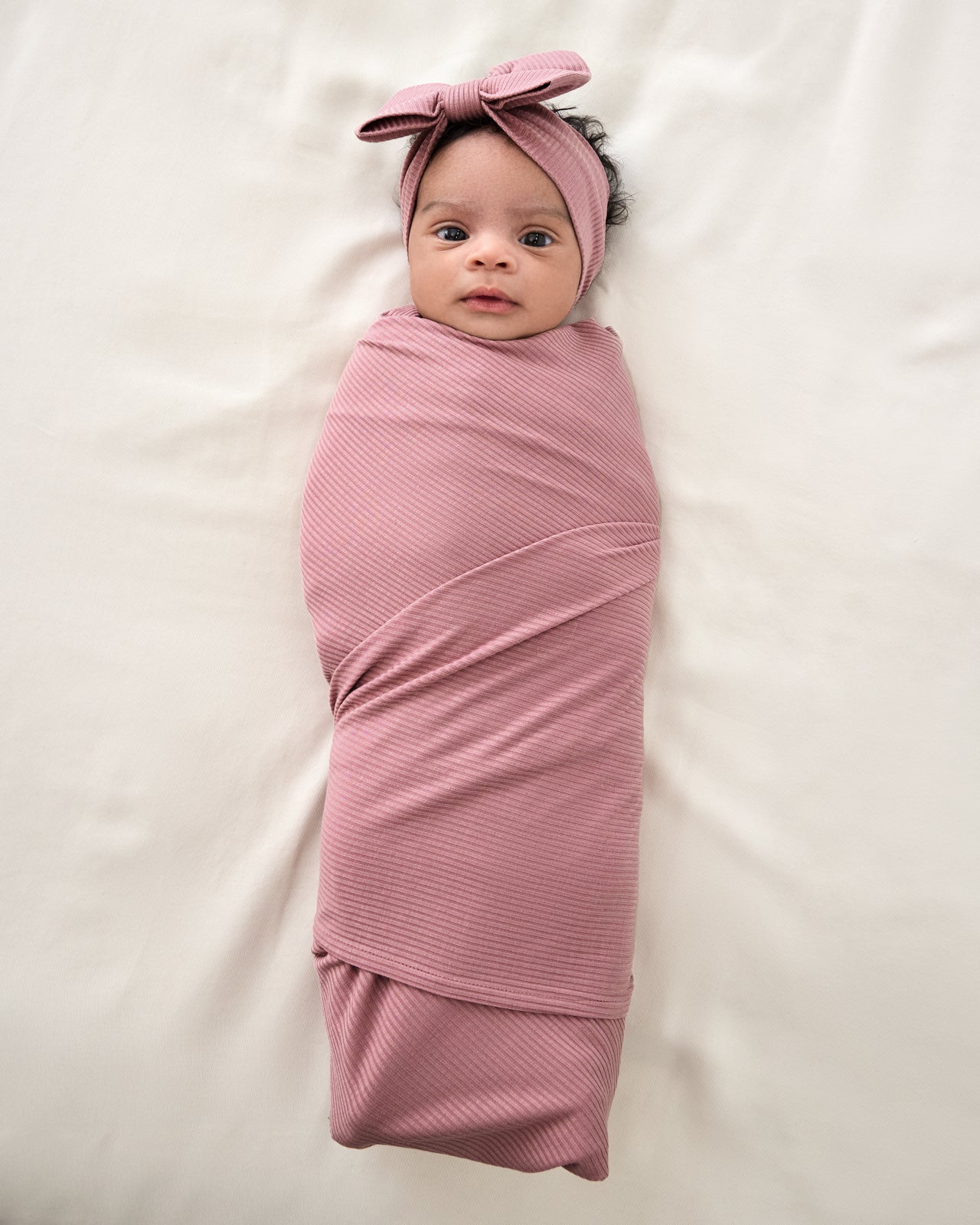 Overhead view of newborn swaddled in the Rosewood Ribbed Swaddle & Luxe Bow Headband Set