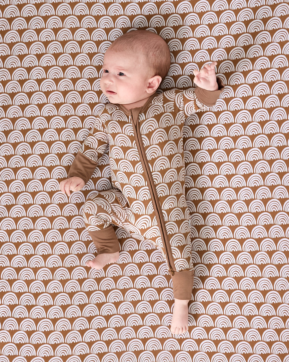 Baby laying on the Rust Rainbows Fitted Crib Sheet wearing a matching zippy