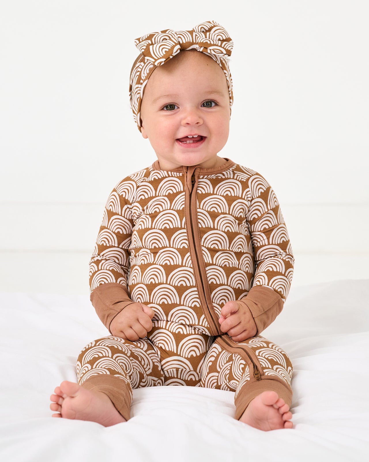 Sitting baby wearing the Rust Rainbows Zippy with a coordinating bow
