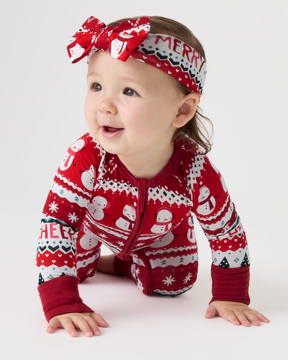 A baby crawling on a light studio floor wearing a red, white, and green Christmas-themed zip-up one-piece pajama with snowman patterns, along with a matching patterned headband.