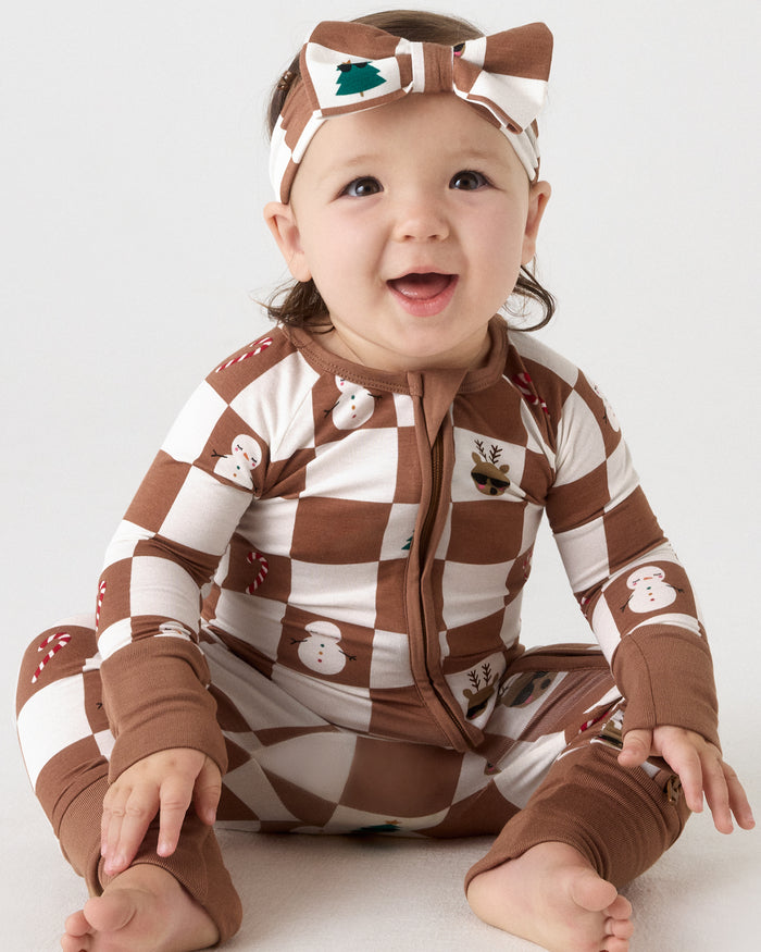 A baby wearing a brown and white checkered zip-up onesie with snowman and reindeer prints, and a matching headband, sitting against a light gray background.