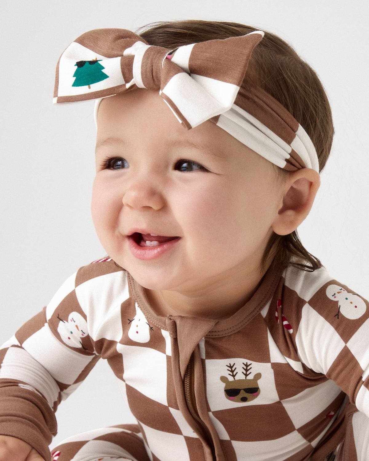A baby wearing a brown and white checkered zip-up onesie with holiday-themed icons and a matching brown and white checkered bow headband, photographed against a light gray studio background.