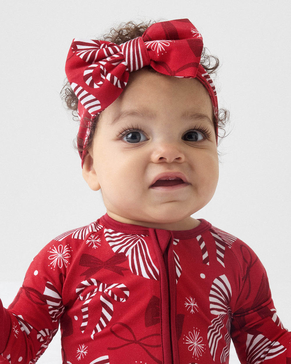 A close-up of a baby wearing a red and white holiday-themed zip-up one-piece pajama and a matching red bow headband with holiday ribbon and snowflake patterns, set against a plain light gray background.