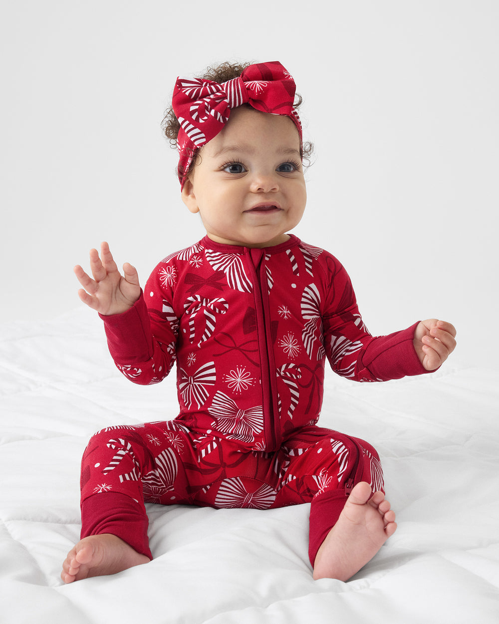 A baby sitting on a white fabric surface wearing a red long-sleeve zip-front one-piece pajama with a white holiday pattern, plus a matching red bow headband.