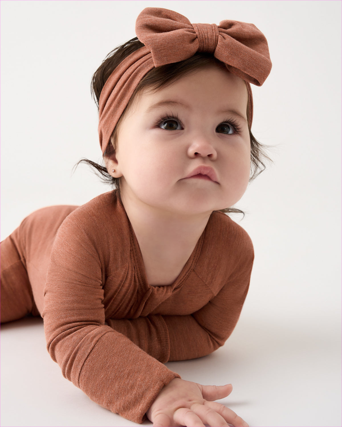 A baby wearing a matching rust-brown long-sleeve bodysuit and large bow headband, lying on a white studio background.