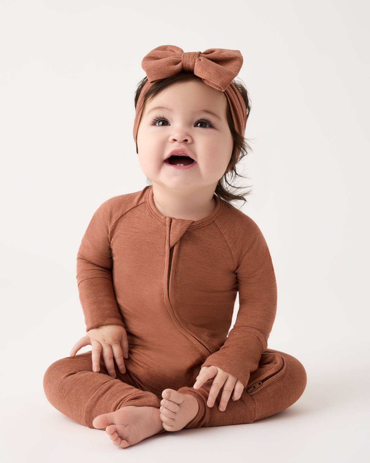 A baby sitting cross-legged wearing a rust-brown long-sleeve zip-front bodysuit and matching bow headband against a plain white studio background.