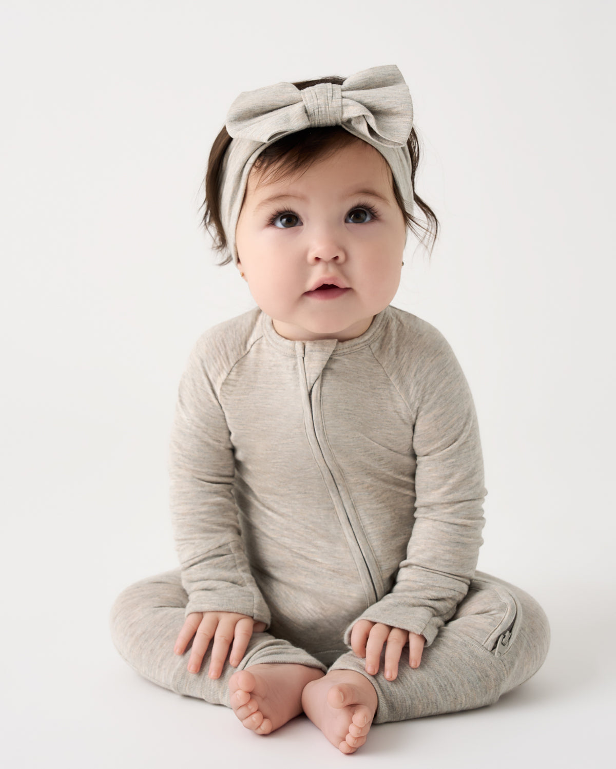 A baby sitting cross-legged wearing a light taupe heathered long-sleeve zip-front footed one-piece and a matching bow headband, photographed against a seamless white studio background.